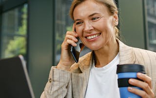 Woman on a business call on mobile phone looking at laptop holding a coffee
