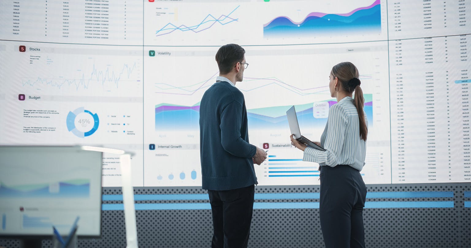 Business man and woman discussing UC analytics in front of a large screen full of statistics