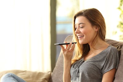 Voss Give Teams a Voice image - Girl using a smart phone voice recognition on line sitting on a sofa in the living room at home with a warm light and a window in the background