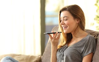 Voss Give Teams a Voice image - Girl using a smart phone voice recognition on line sitting on a sofa in the living room at home with a warm light and a window in the background